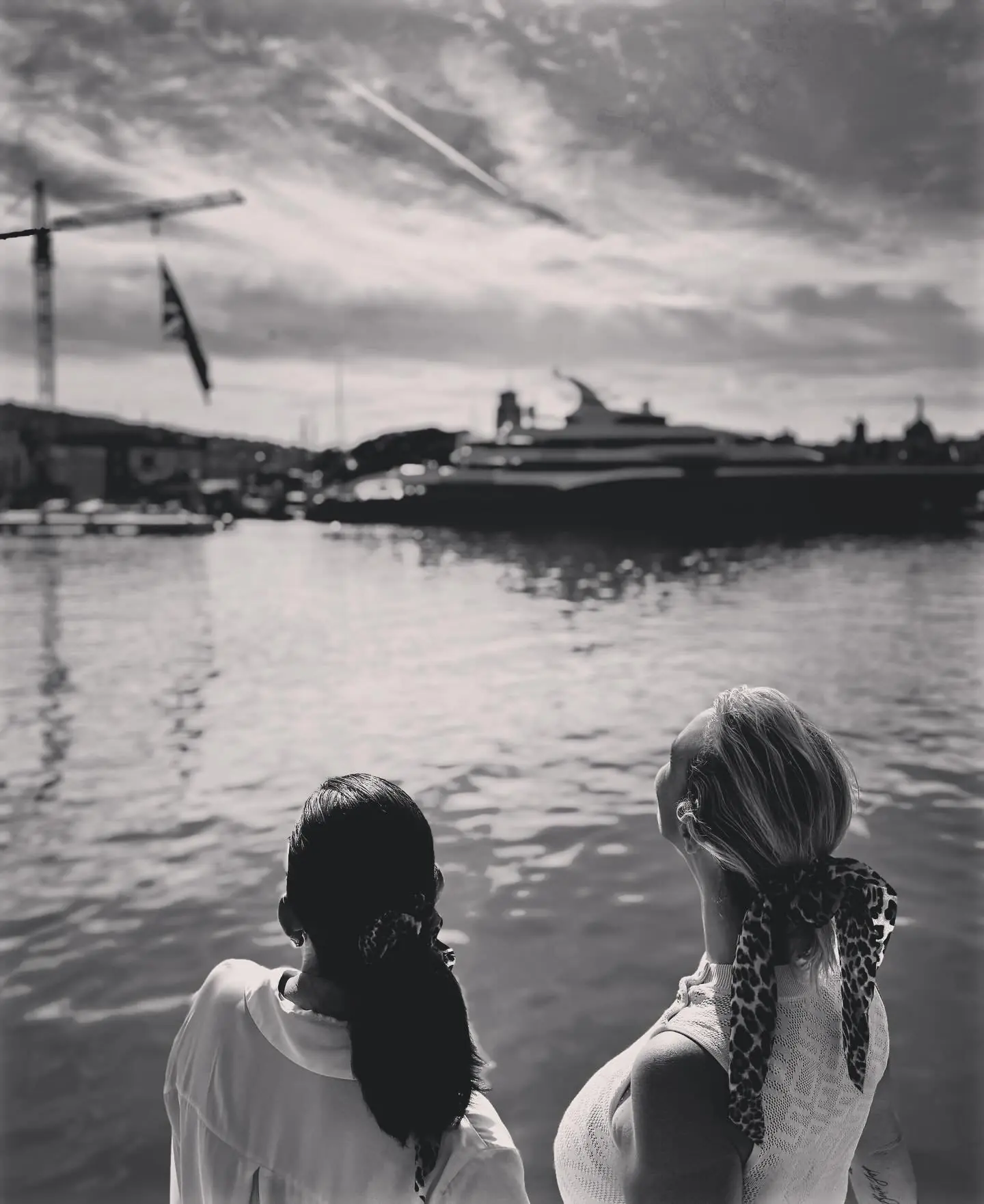 Two women seen from behind looking out to sea from the harbour, in black and white