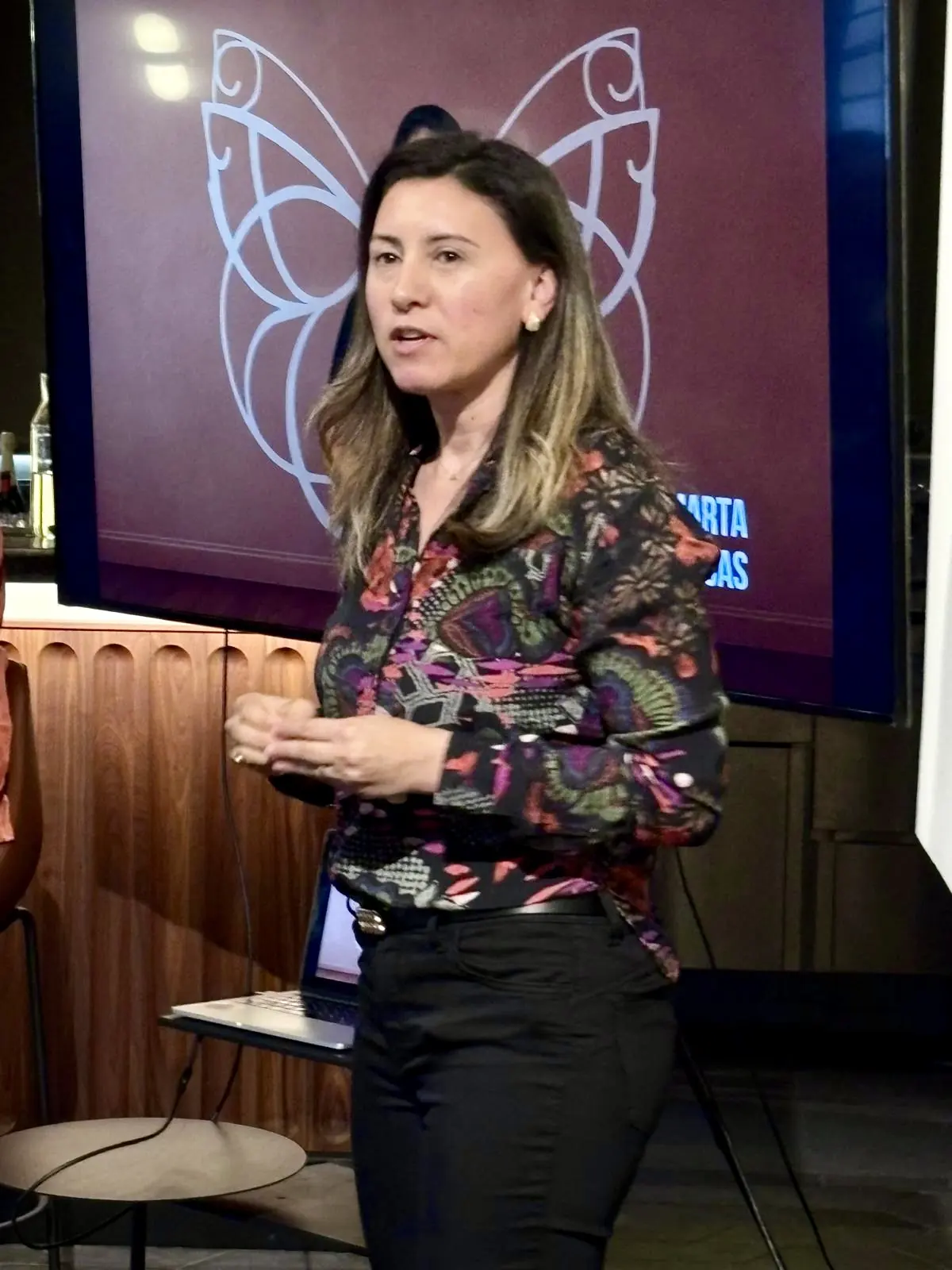 A speaker addresses the audience in front of a screen showing the IKONIKAS butterfly